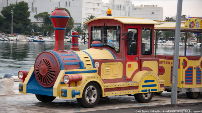 Bright red, yellow, and blue tourist road train styled like a locomotive beside Zadar’s marina.