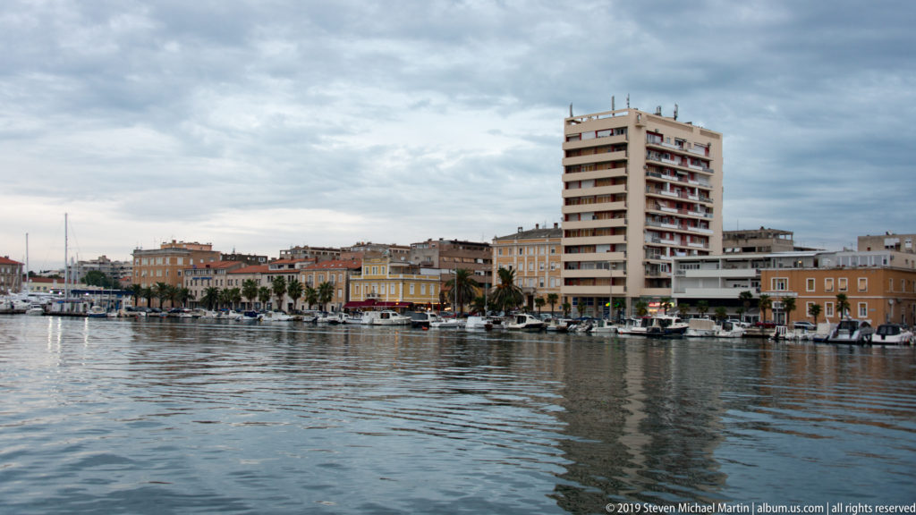 Harbor view of Zadar with moored boats, low historic waterfront buildings, and a tall modern apartment block.
