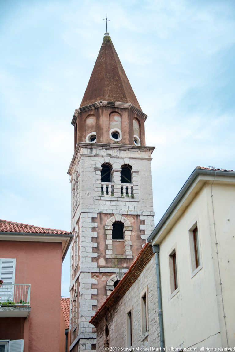 Square pale-stone bell tower with arched openings, reddish octagonal upper stage, pyramidal roof, and cross.