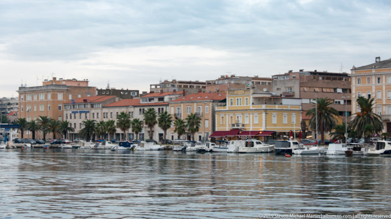 Zadar harbor with palm-lined waterfront buildings and numerous small boats moored along the quay.