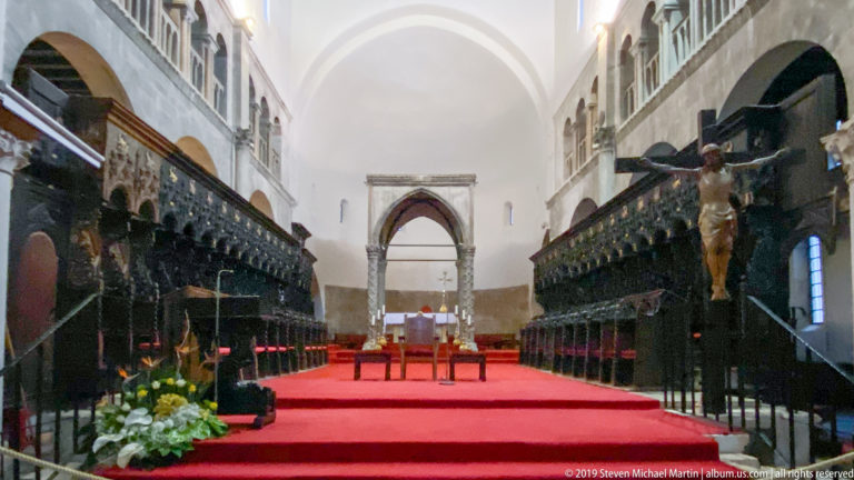 Carved wooden choir stalls flank the red-carpeted presbytery of St. Anastasia Cathedral, with a crucifix at right.