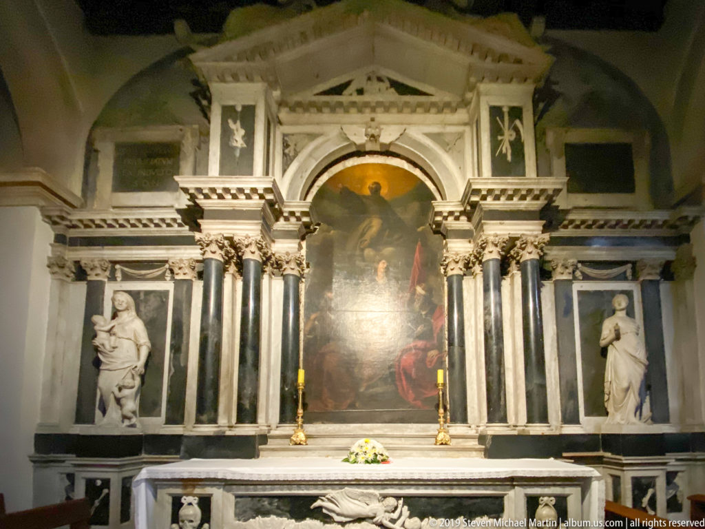 Ornate marble church altar with paired columns, saint statues, central painting, candles, flowers, and a relief-decorated altar table.