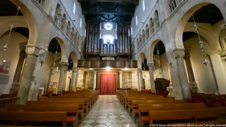 Interior nave of St. Anastasia Cathedral with stone arcades, timber roof, wooden pews, and organ loft.