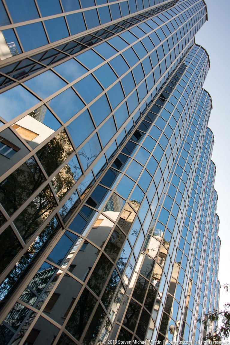 Extreme low-angle view up a curving blue-glass office tower with gridded mullions and reflections of nearby trees and buildings.