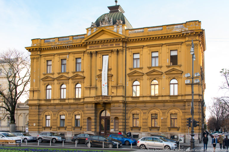 Ochre neo-Renaissance institutional building with central copper-roofed pavilion, tall windows, banners, and cars along the street.