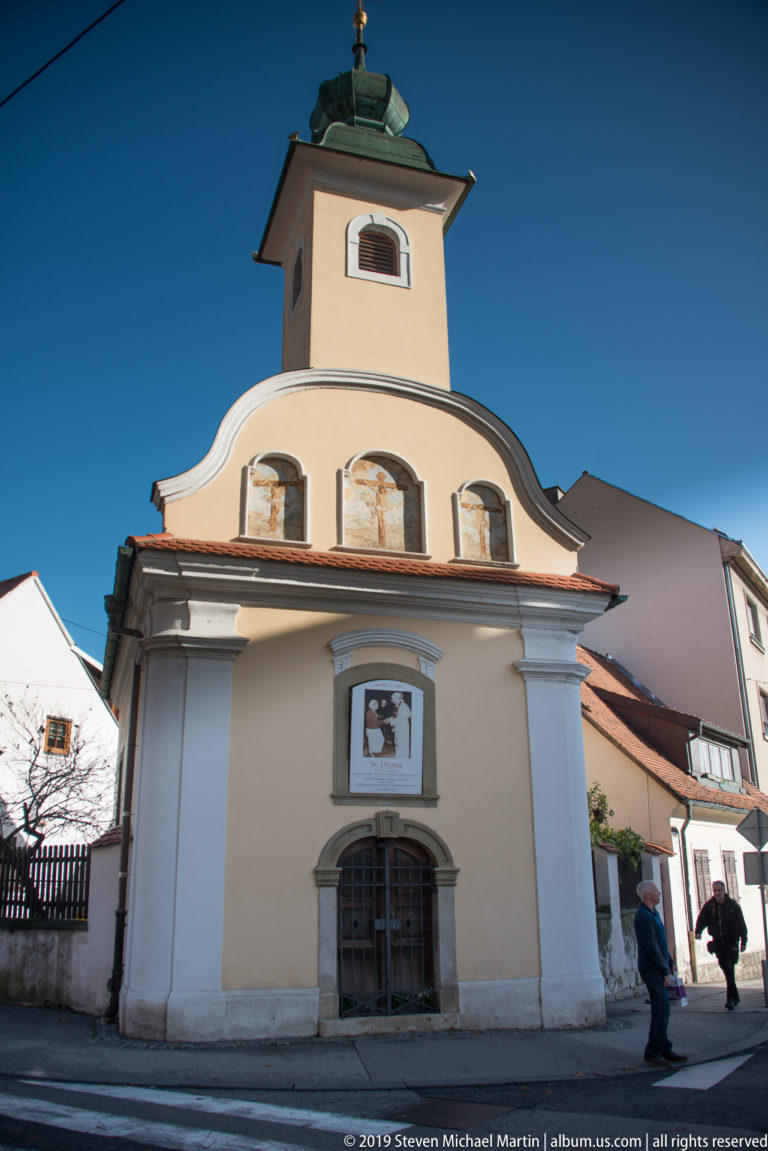 Small pale chapel with curved Baroque-style gable, square bell stage, three arched panels, and a central entrance below.