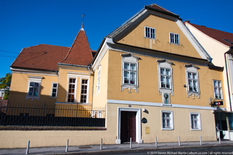 Yellow historic house with steep red-tiled roof, gabled front, white-trimmed windows, doorway, and attached lower wing.