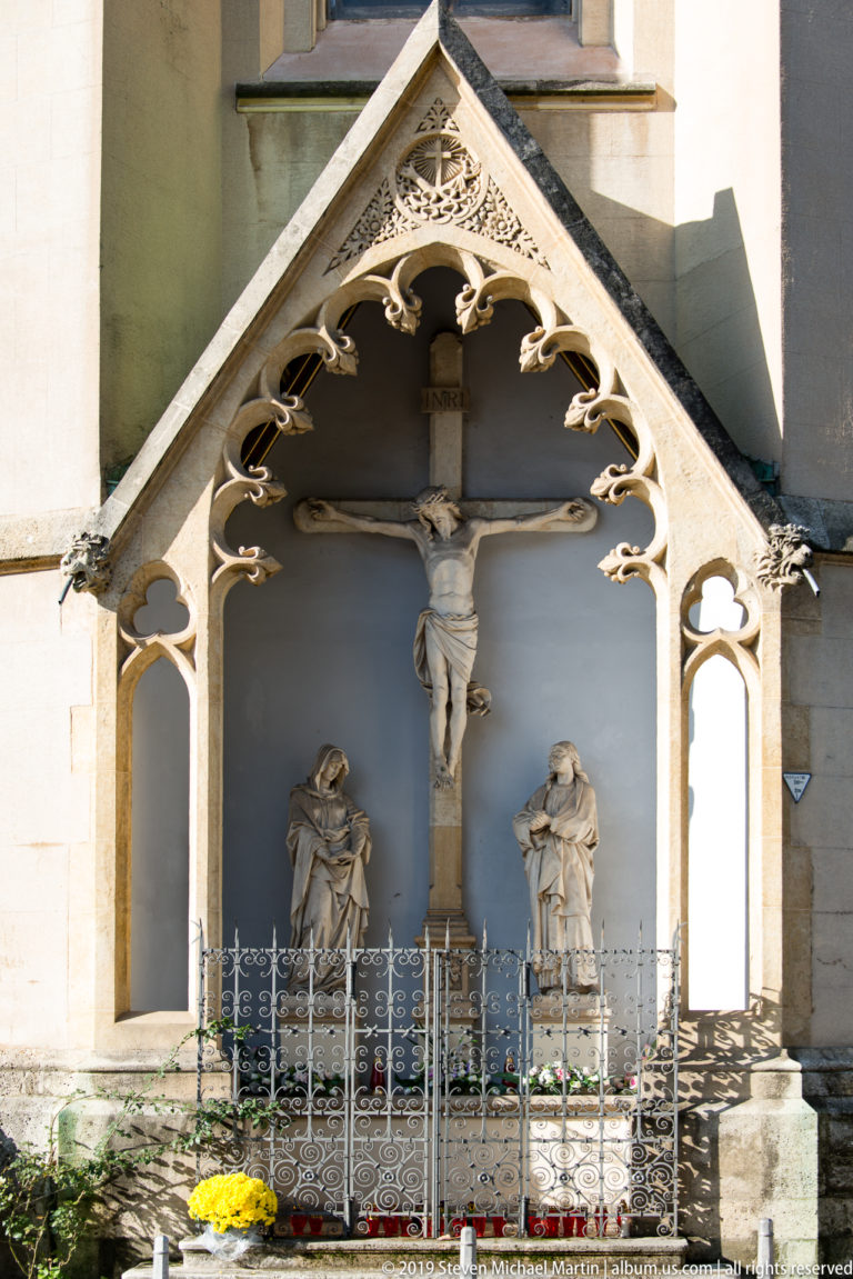 Crucifixion shrine beneath a pointed Gothic canopy, with Christ on the cross, two flanking figures, candles, and flowers.