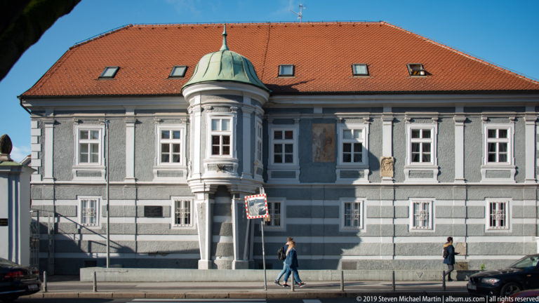 Gray historic building with striped façade, red-tiled roof, rounded projecting bay with green cap, and pedestrians outside.