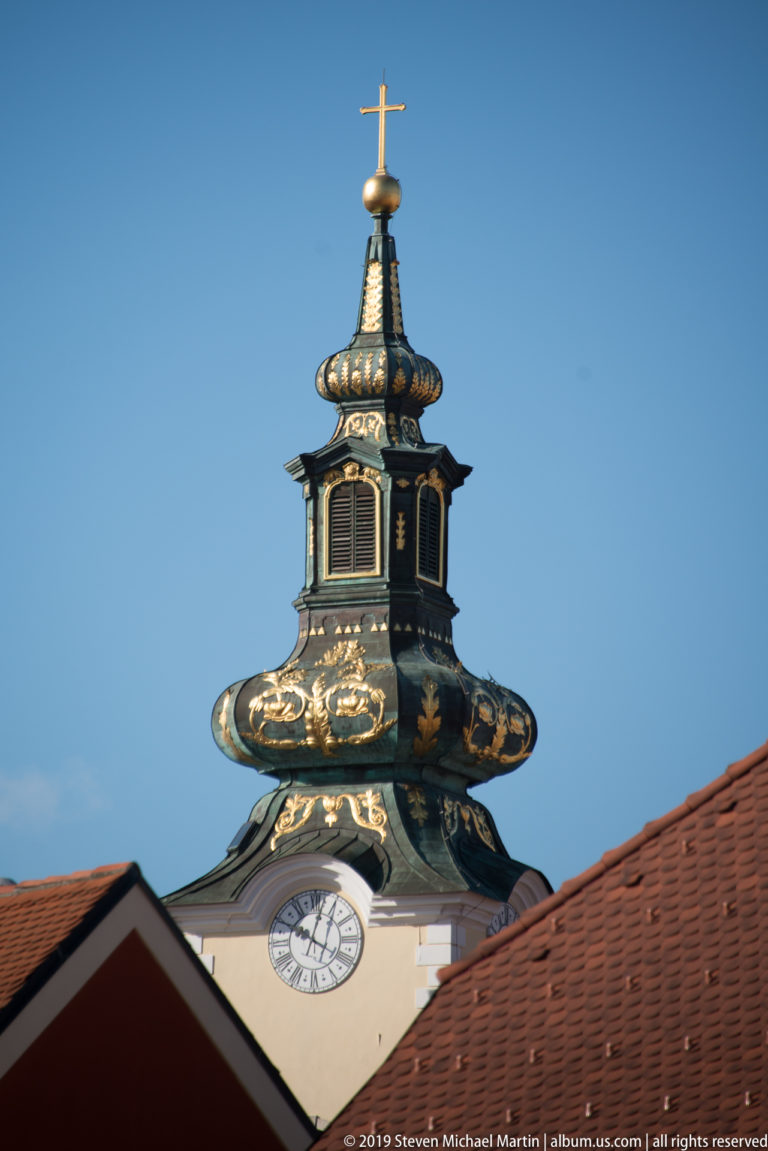Ornate dark metal clock tower with gilded decoration, curved roof forms, and a cross above red-tiled roofs.