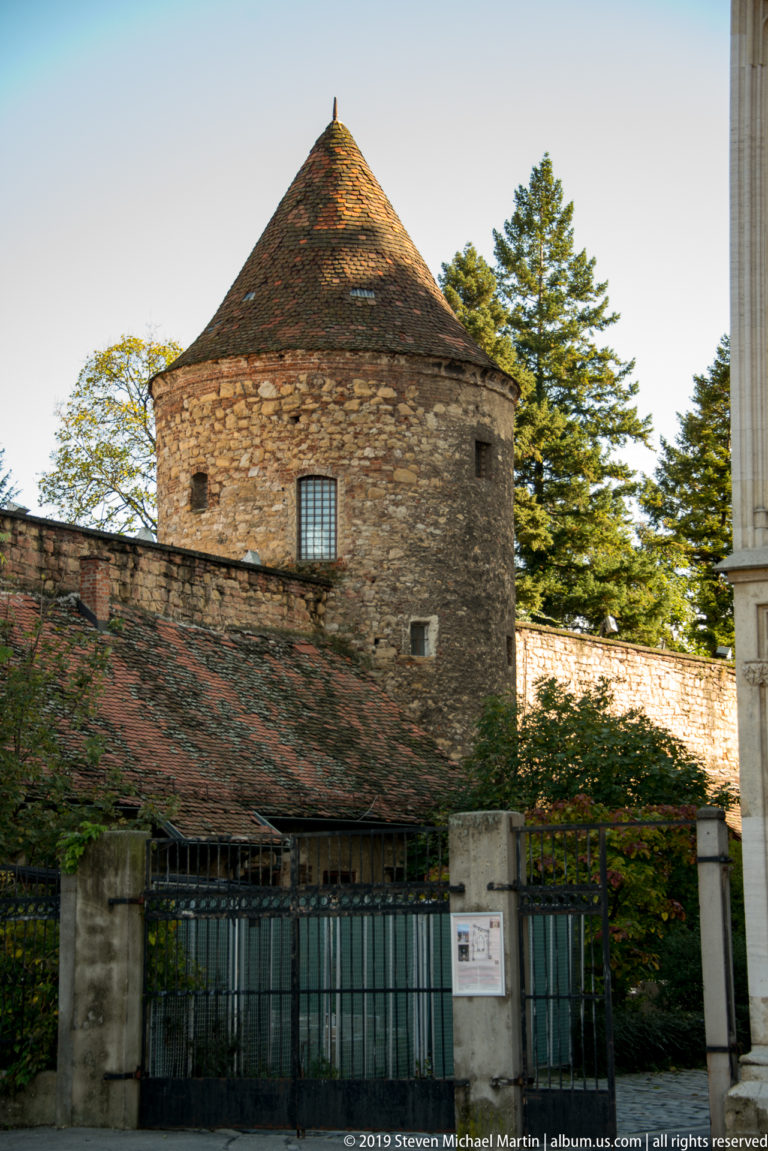 Round stone defensive tower with small openings and a conical tiled roof behind a wall and iron gate in Zagreb.