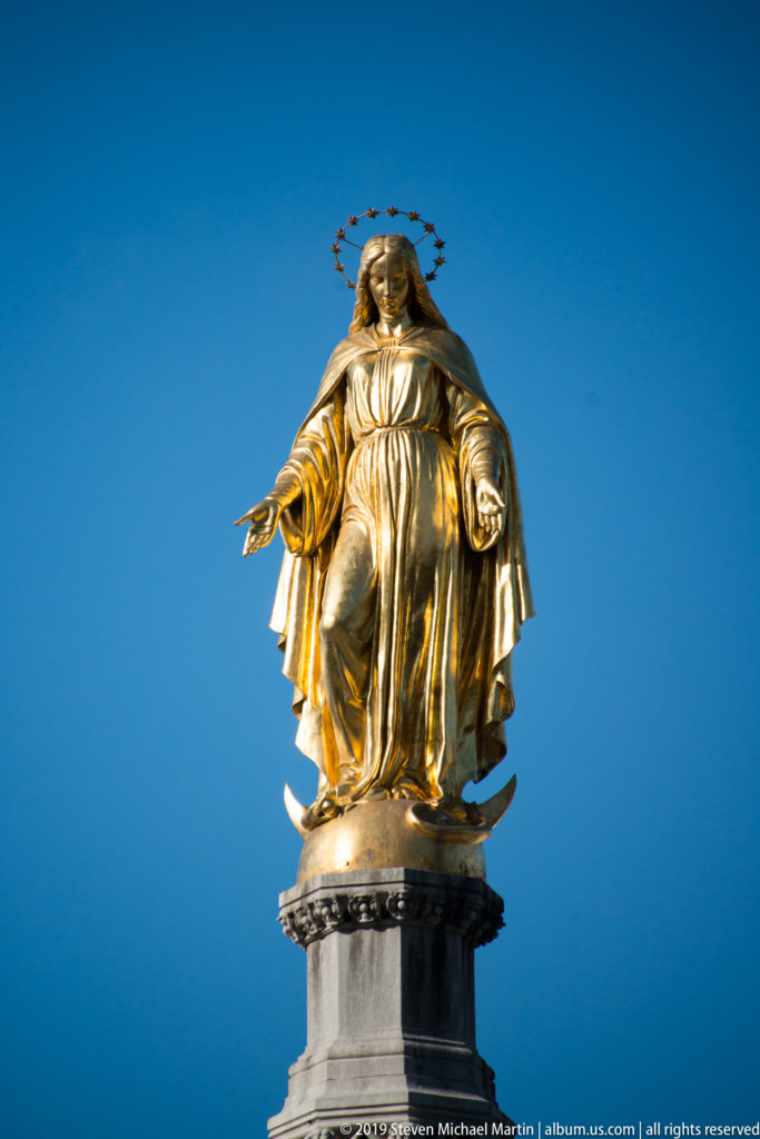 Close-up of gilded Virgin Mary with crown, star halo, flowing robes, and open hands atop a dark column capital.