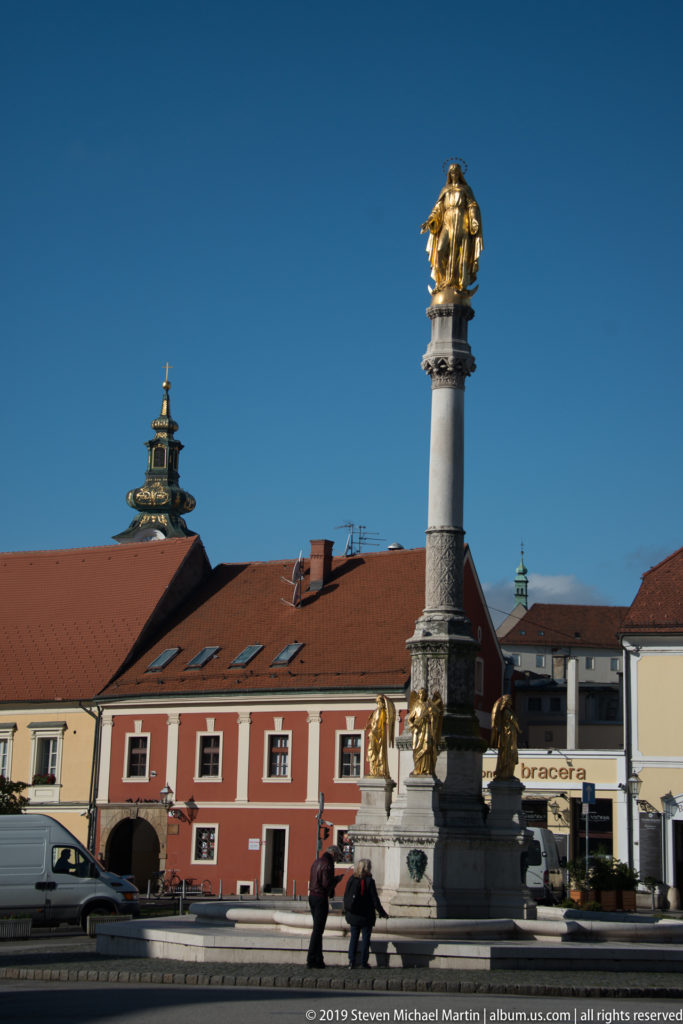 Tall Marian column with a gilded figure on top and smaller gold figures at the base, surrounded by historic Zagreb buildings.