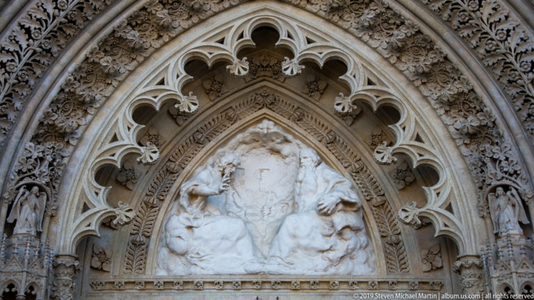 Close view of a pointed cathedral tympanum filled with pale stone relief figures, Gothic tracery, foliage, and jamb sculpture.
