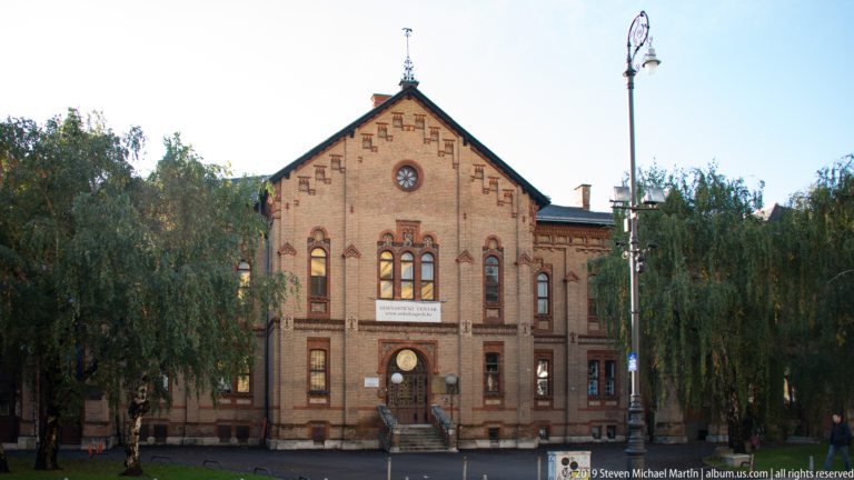 Symmetrical brick historic building with central gable, clock, arched windows, entrance banner, and trees on both sides.