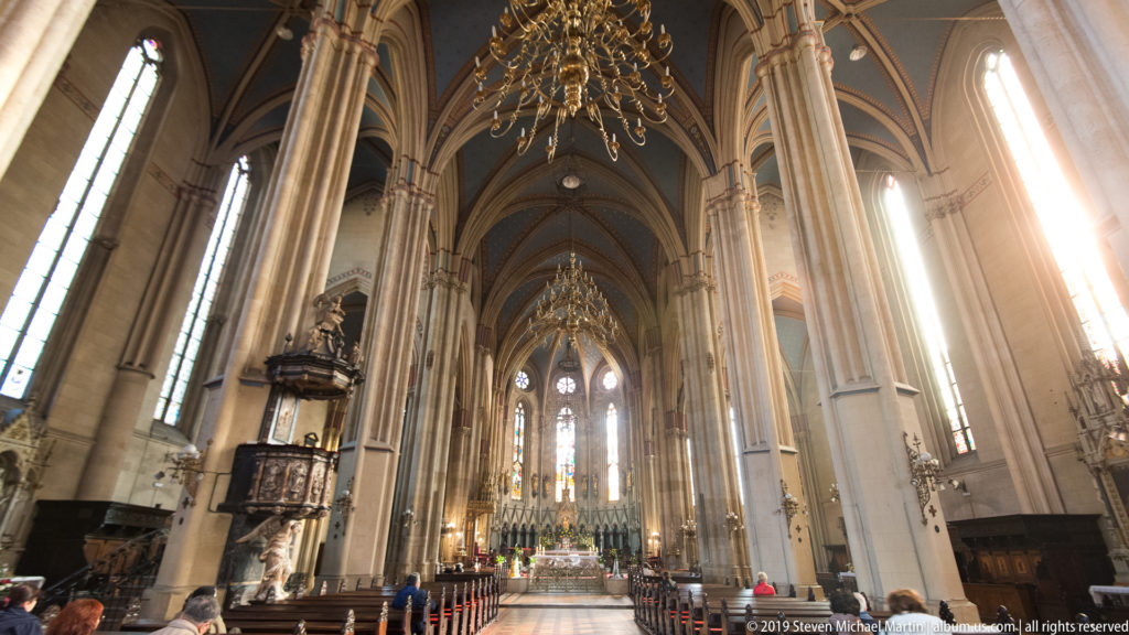 Sunlit nave of Zagreb Cathedral with tall piers, ribbed vaults, chandelier, pulpit, and distant altar.