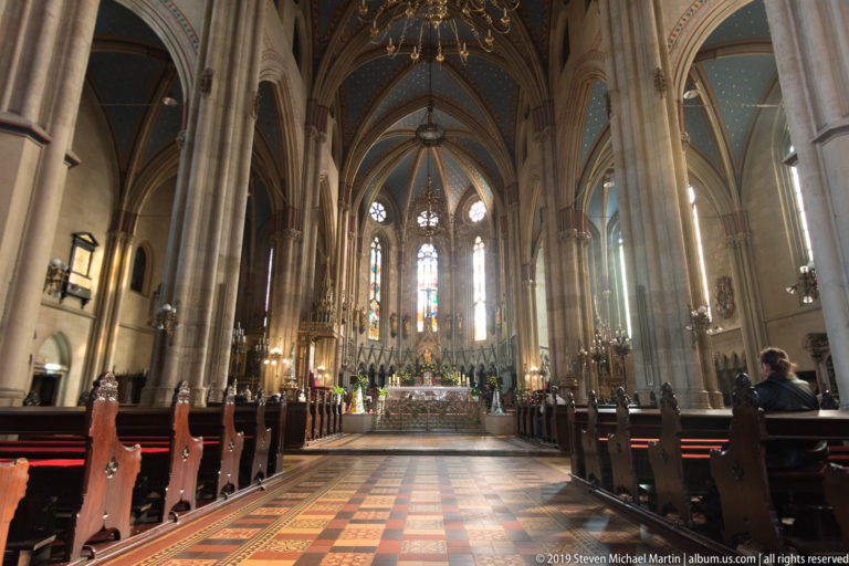 Symmetrical central aisle of Zagreb Cathedral leading past pews and columns toward the illuminated high altar and stained glass.