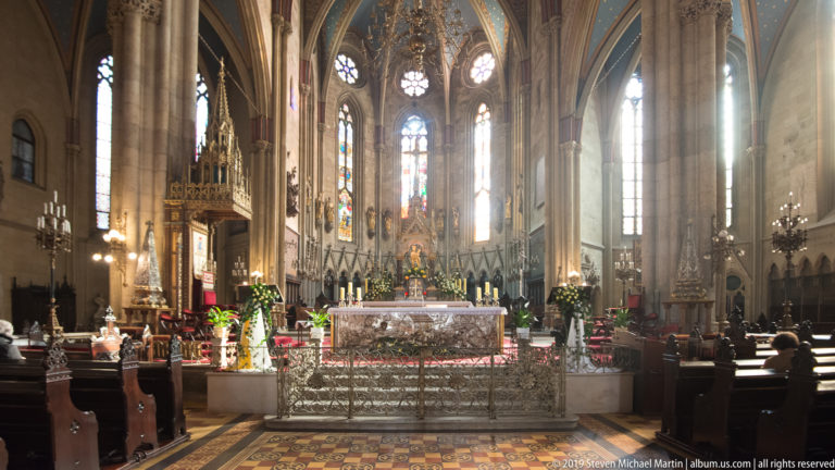 Central nave of Zagreb Cathedral facing the altar, with ribbed vaults, stained glass, columns, pews, and patterned floor.