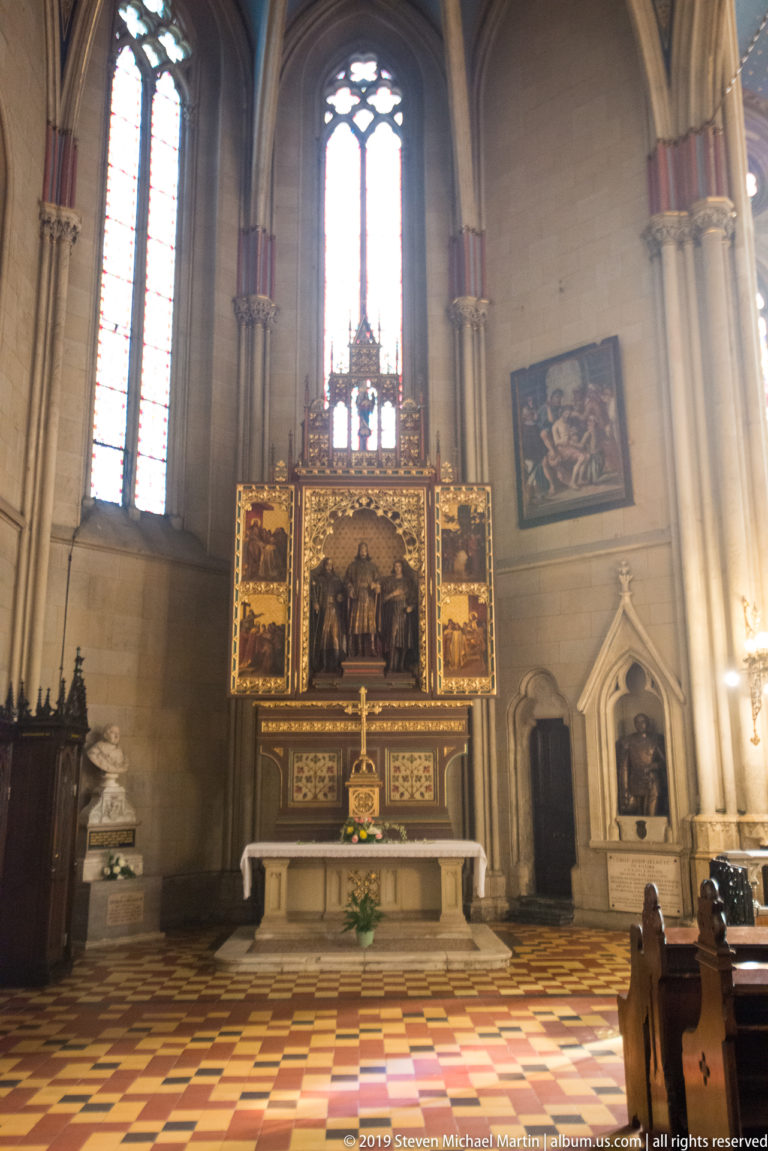 Gilded side chapel altar beneath a tall stained-glass window, with painted panels, nearby artwork, and patterned floor tiles.