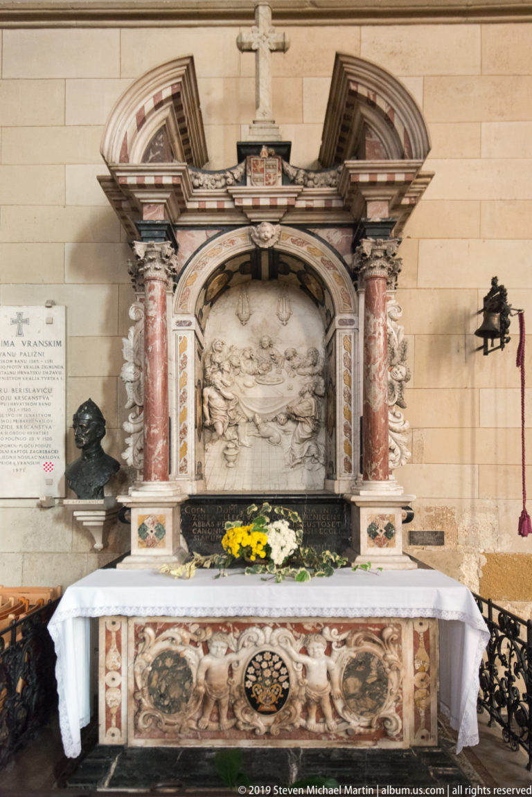 Polychrome side altar inside Zagreb Cathedral with red columns, central relief, gilded details, flowers, and white altar cloth.