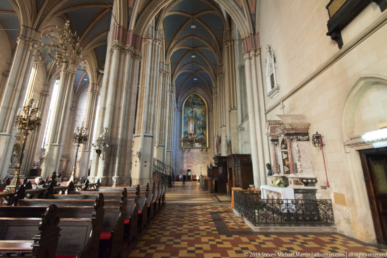 Long side aisle in Zagreb Cathedral with ribbed vaults, patterned floor, pews, side altar, and distant painted altarpiece.