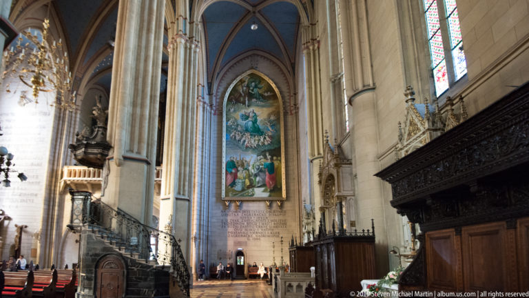 Side aisle of Zagreb Cathedral with tall vaults, large religious painting, inscribed wall, pulpit, and dark carved woodwork.