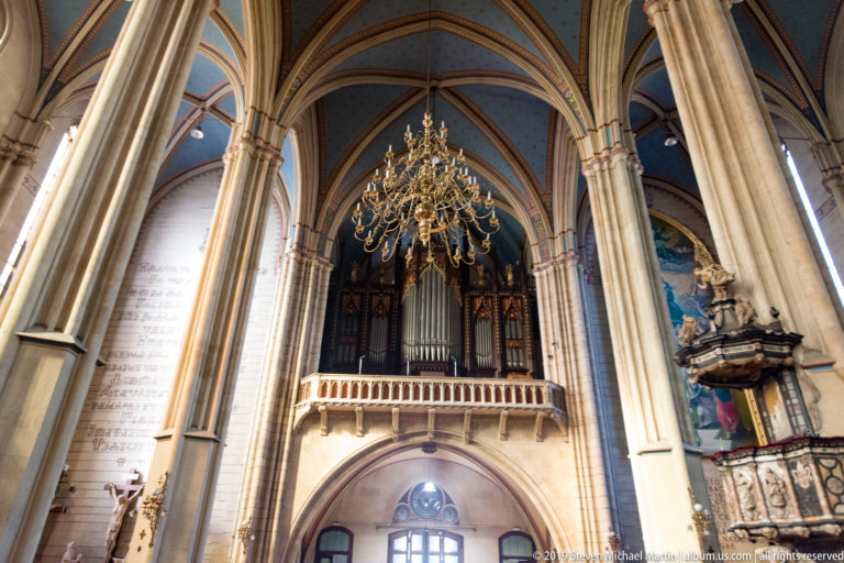 Pipe organ on a gallery inside Zagreb Cathedral, framed by tall columns, ribbed vaults, and a hanging chandelier.