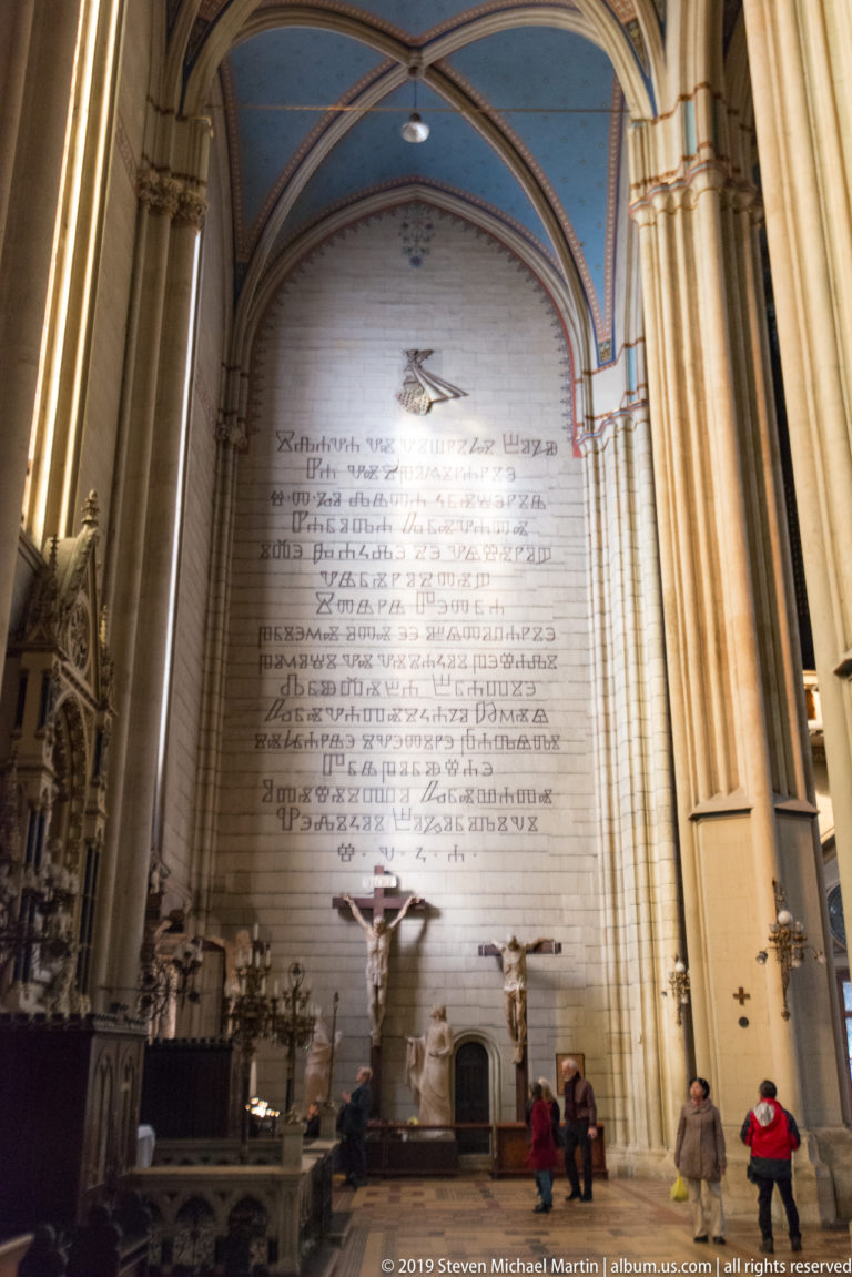 Tall inscribed wall panel inside Zagreb Cathedral beside a crucifix and church furnishings, with visitors below for scale.