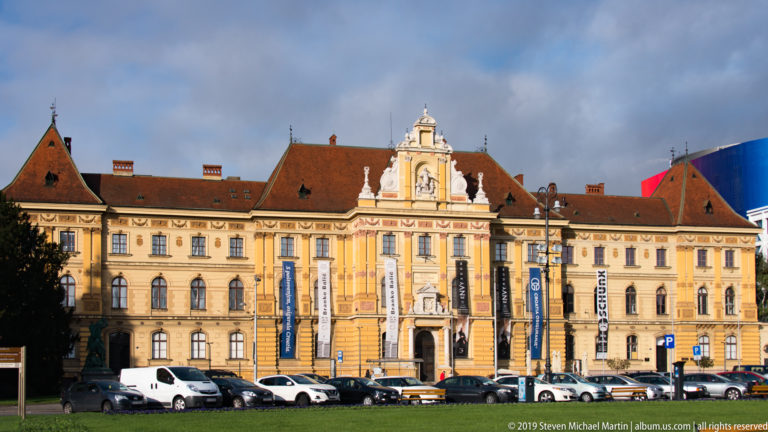 Long ochre museum façade with red tile roofs, ornate central entrance pavilion, banners, and parked cars in Zagreb.