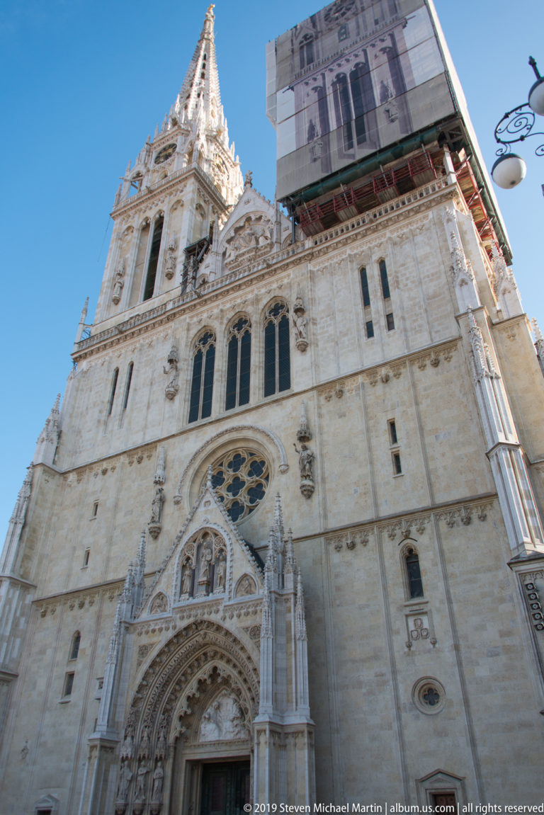 Close low-angle view of Zagreb Cathedral’s west front, with ornate portal, rose window, twin towers, and restoration wrap.