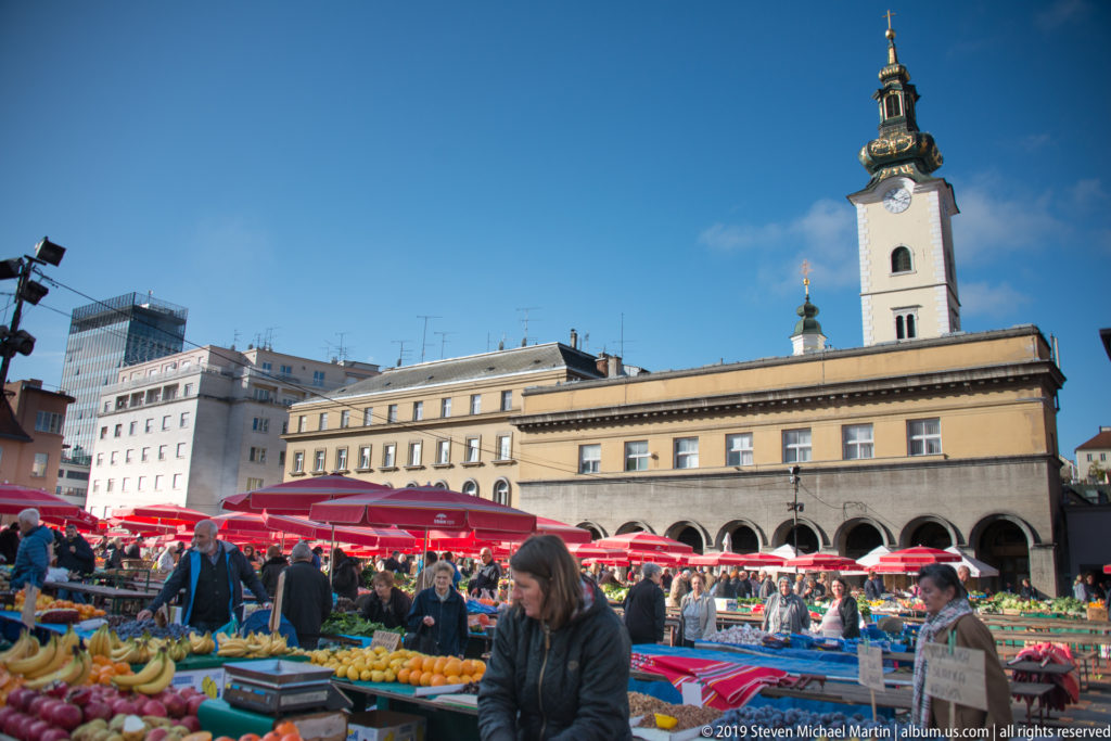 Busy Dolac Market with produce stalls under red umbrellas, shoppers, surrounding buildings, and a church tower behind.