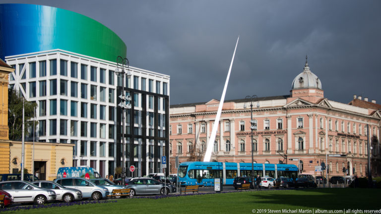 Tall white sculpture, blue tram, modern glass building, and pink historic palace share a Zagreb square beneath dark clouds.