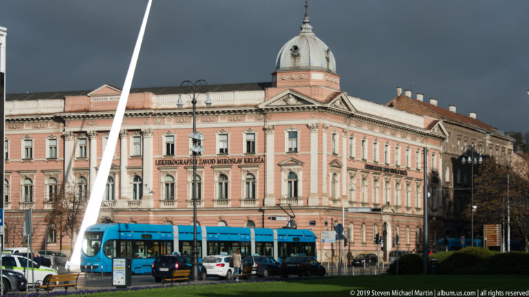 Blue tram passes a pink domed historic institutional building behind a tall white sculpture and landscaped square in Zagreb.