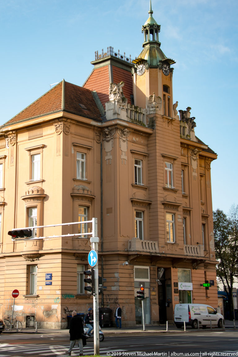 Ochre historic corner building with classical windows, rooftop pavilion, green metal roof, and modern traffic signals.