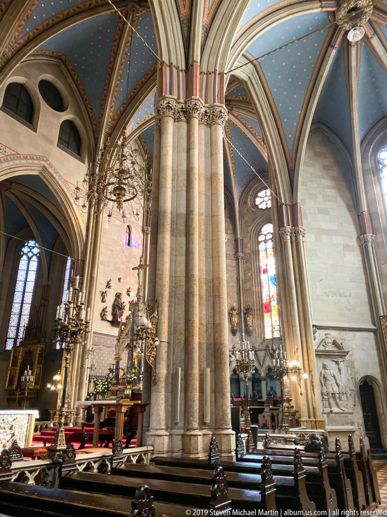 Massive clustered Gothic pier in Zagreb Cathedral with arches, stained glass, side altars, chandeliers, and seating beyond.