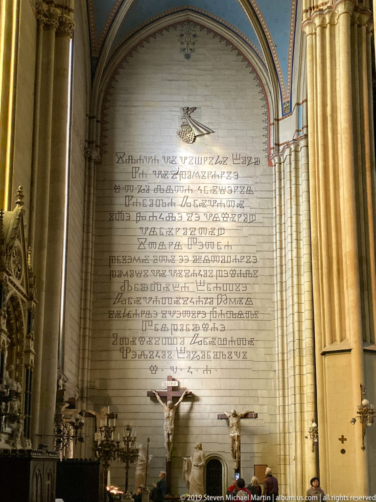 Large multi-line wall inscription above a crucifix and candles inside a Gothic cathedral, framed by tall stone columns.