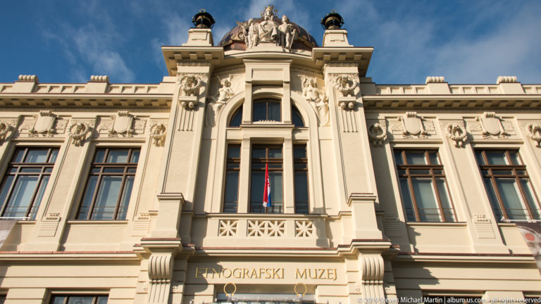 Front façade of Zagreb’s Ethnographic Museum with ETNOGRAFSKI MUZEJ inscription, central arched window, sculptures, and copper roof.
