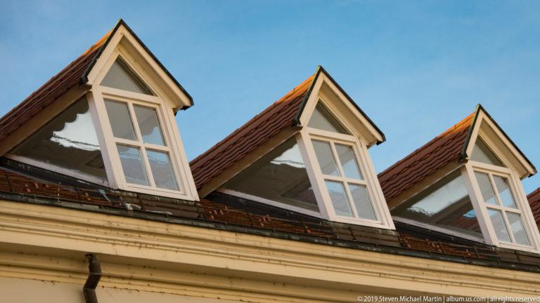 Three cream-painted gabled dormer windows project from a red clay-tiled roof against a blue sky.
