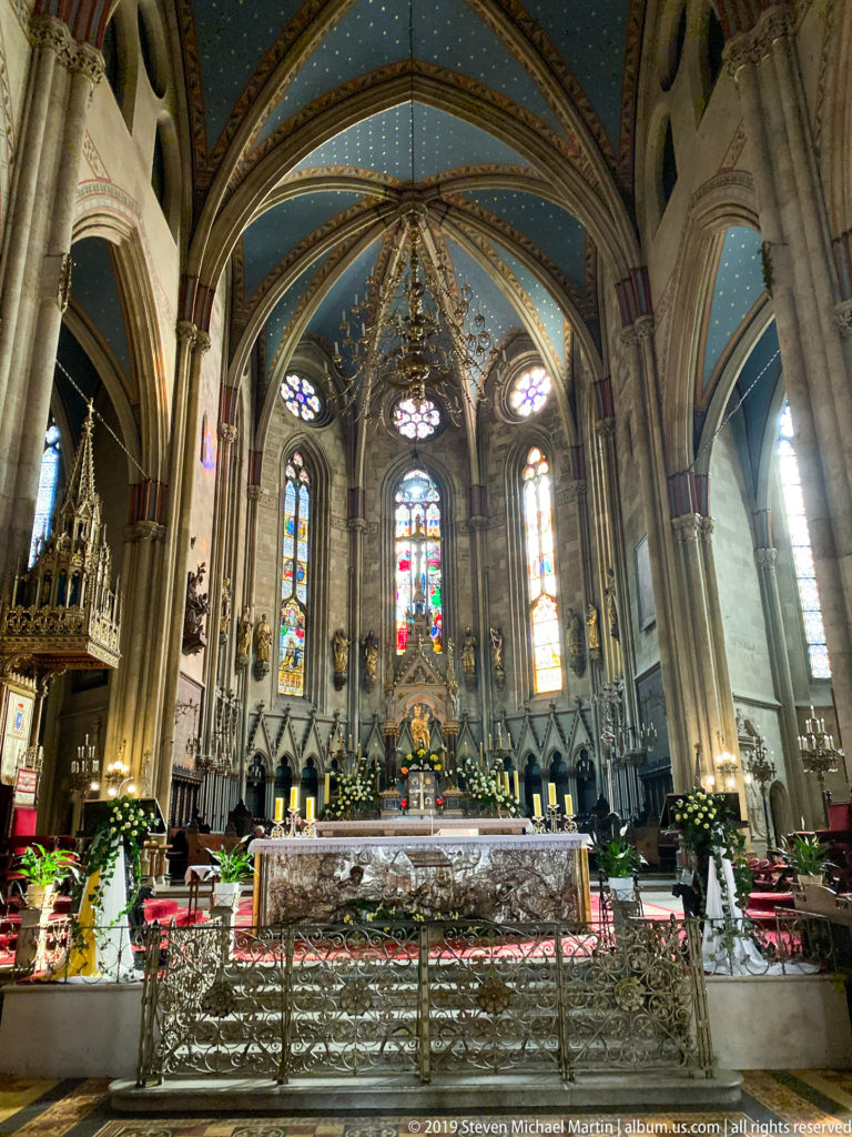 Zagreb Cathedral sanctuary with altar, flower arrangements, tall stained-glass windows, clustered columns, and blue ribbed vaulting.