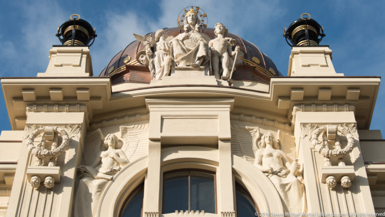 Upper façade of Zagreb’s Ethnographic Museum with copper dome, seated rooftop figure, arched window, wreaths, and flanking sculptures.