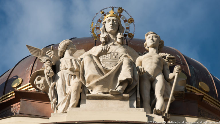 Seated female allegorical sculpture with smaller figures before the copper dome of Zagreb’s Ethnographic Museum.
