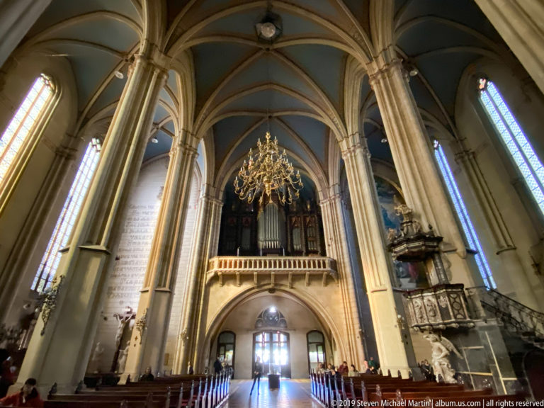 Rearward cathedral nave view with organ gallery, chandelier, clustered piers, blue ribbed vaults, stained glass, and bright entrance doors.