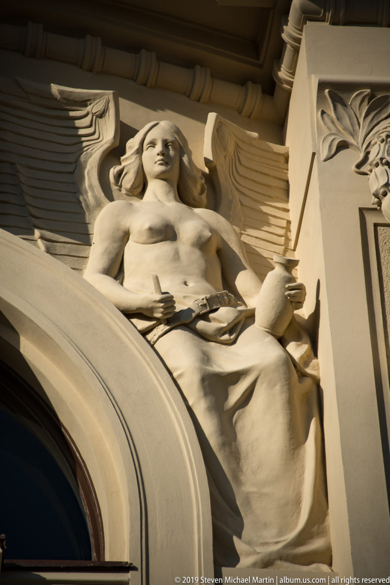 Pale carved female figure with wings and drapery integrated into the curve of an arched historic façade.