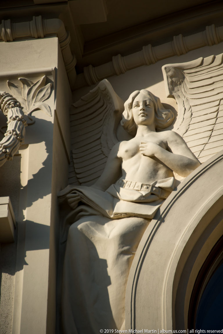 Winged female architectural figure with flowing drapery carved beside the curve of an arched window.