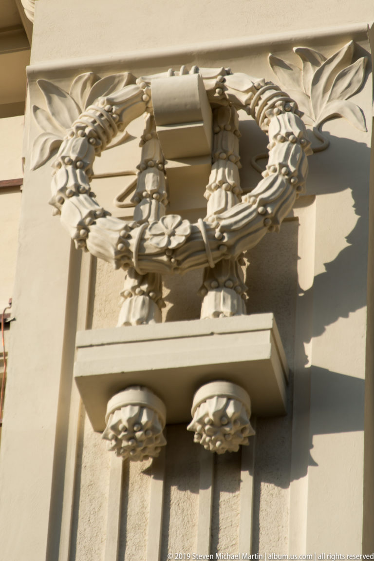 Close view of a sculpted laurel wreath with tied ribbon beneath a projecting pale stone entablature.