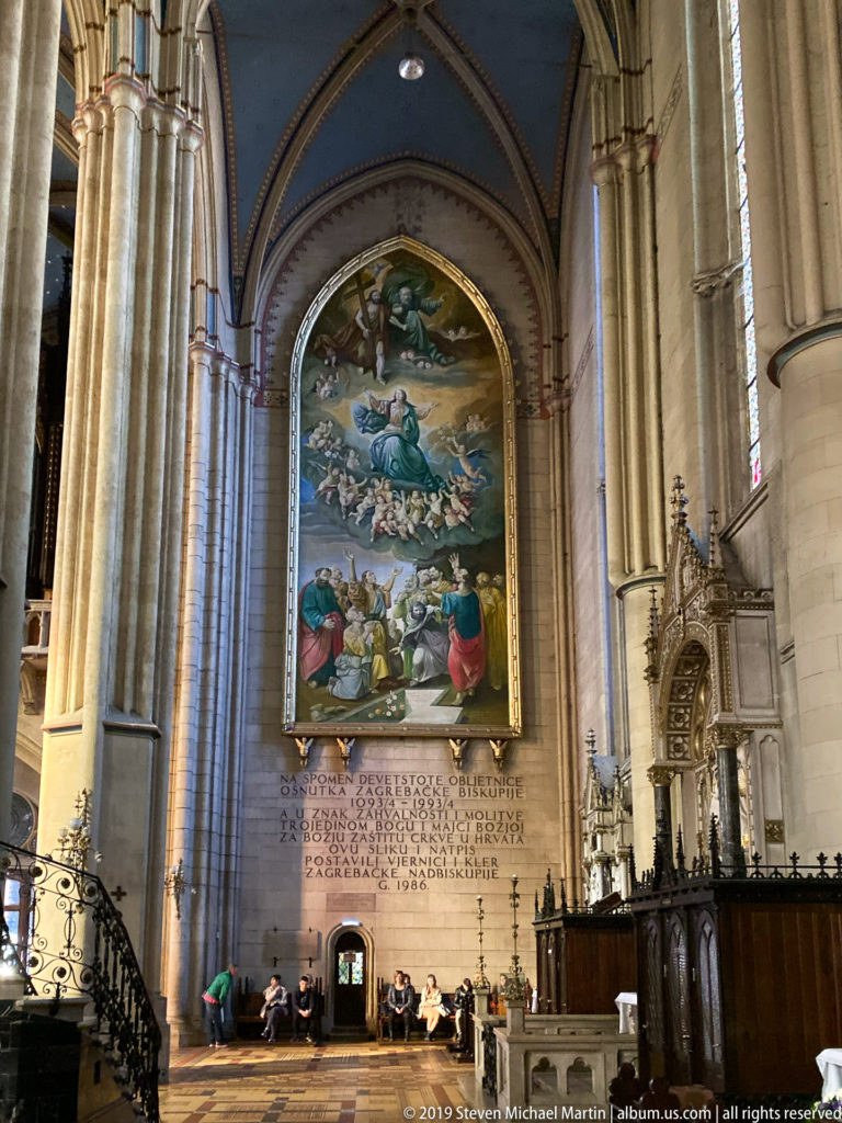 Large framed religious painting above a long wall inscription in a Gothic cathedral aisle with tall stone columns.