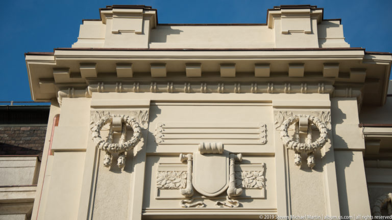 Cream historic façade detail with blank shield cartouche, two sculpted wreaths, foliage, and heavy cornice shadows.