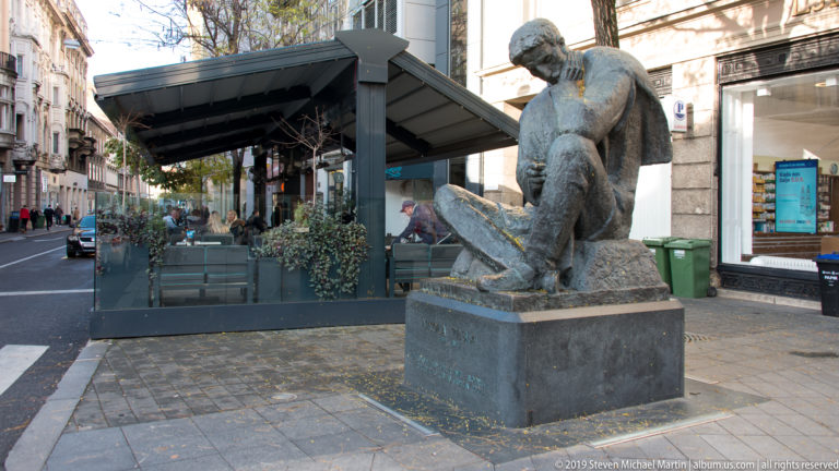 Seated bronze male figure on a stone pedestal beside a covered café terrace and busy pedestrian street in Zagreb.