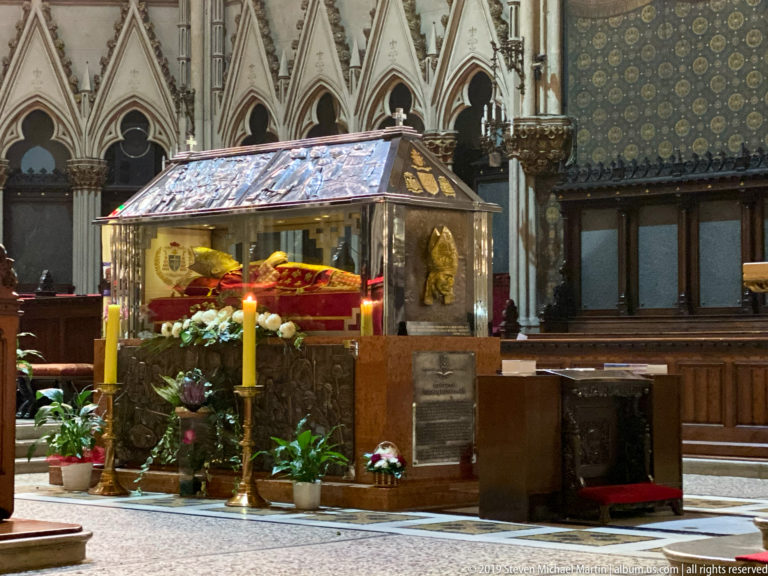 Glass reliquary shrine with red interior, flowers, candles, and a recumbent sacred figure inside Zagreb Cathedral.