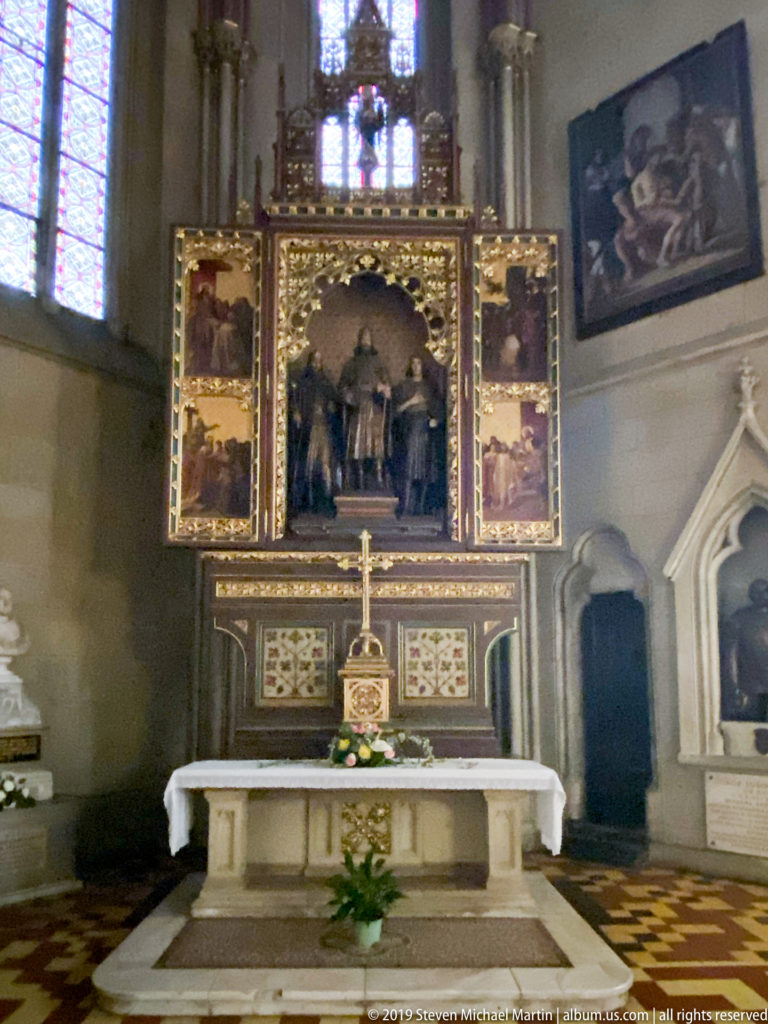 Stone side altar beneath a gilded painted retable, with stained glass and Gothic openings visible around the chapel.